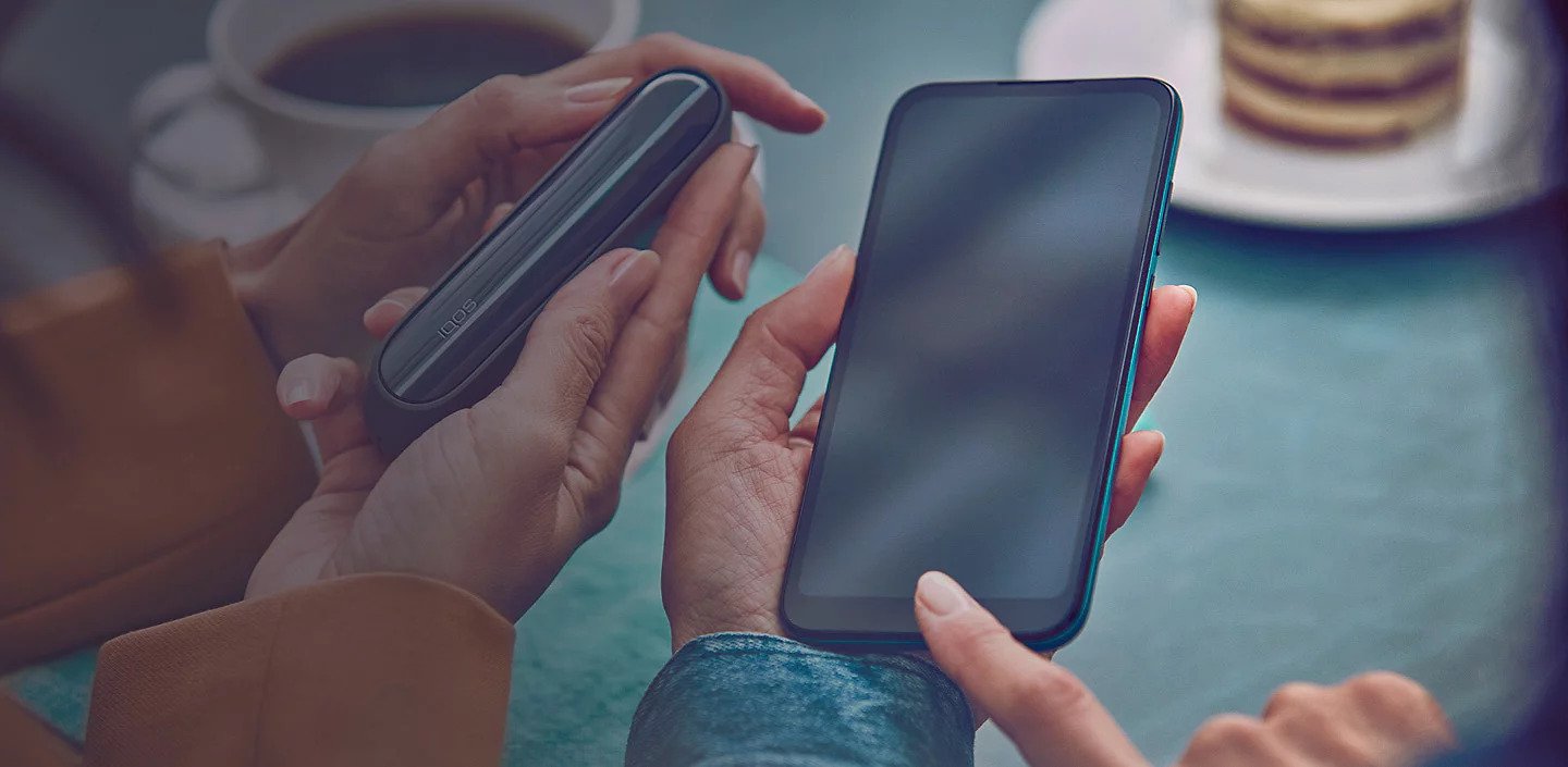 An IQOS device held by one person and a smartphone held by another person.